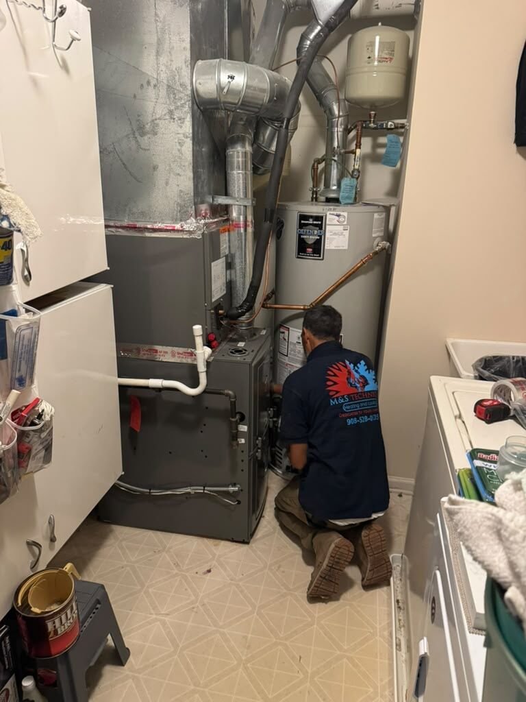 An M&S Technician team member in a branded shirt servicing a gas furnace beside a water heater in a home utility closet